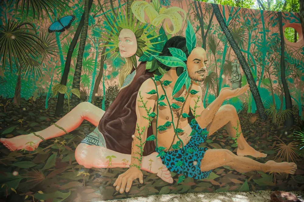 Mural of two seated figures crowned with leaves, back to back inside a painted jungle with a blue butterfly, at Holistika Art Walk in Tulum.