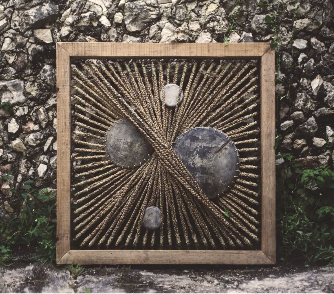A square wooden frame woven with radiating rope and set with smooth river stones, mounted against a rough stone wall.