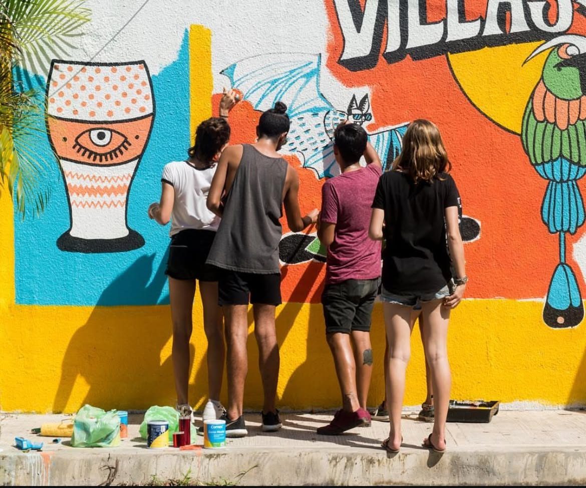 Four artists painting a large street mural of a glass, an eye and a tropical bird on a wall in Tulum, Mexico.