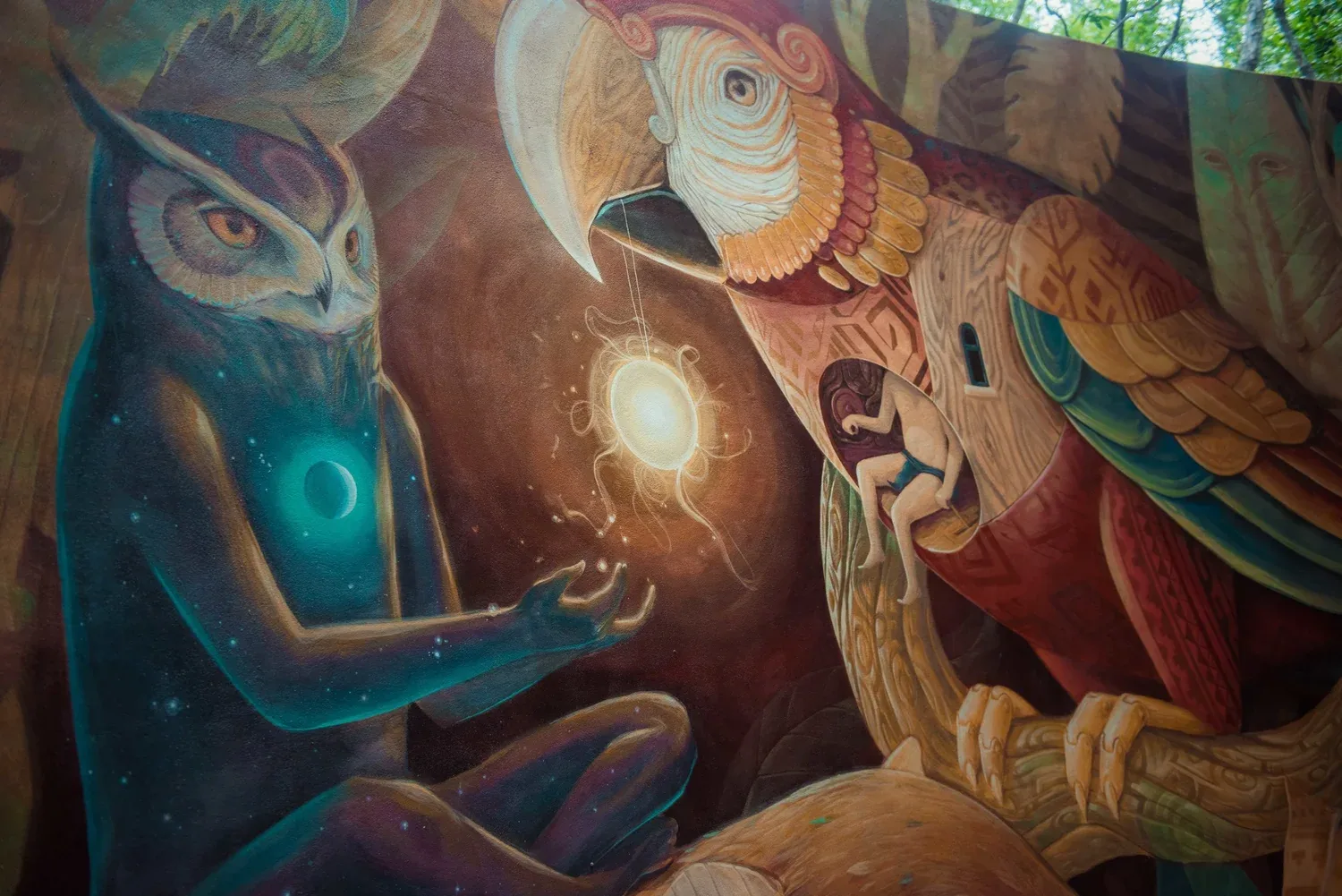 Mural of a blue owl figure holding a glowing sun in front of a large parrot sheltering a sleeping human figure, painted on a curved wall in the Tulum jungle.