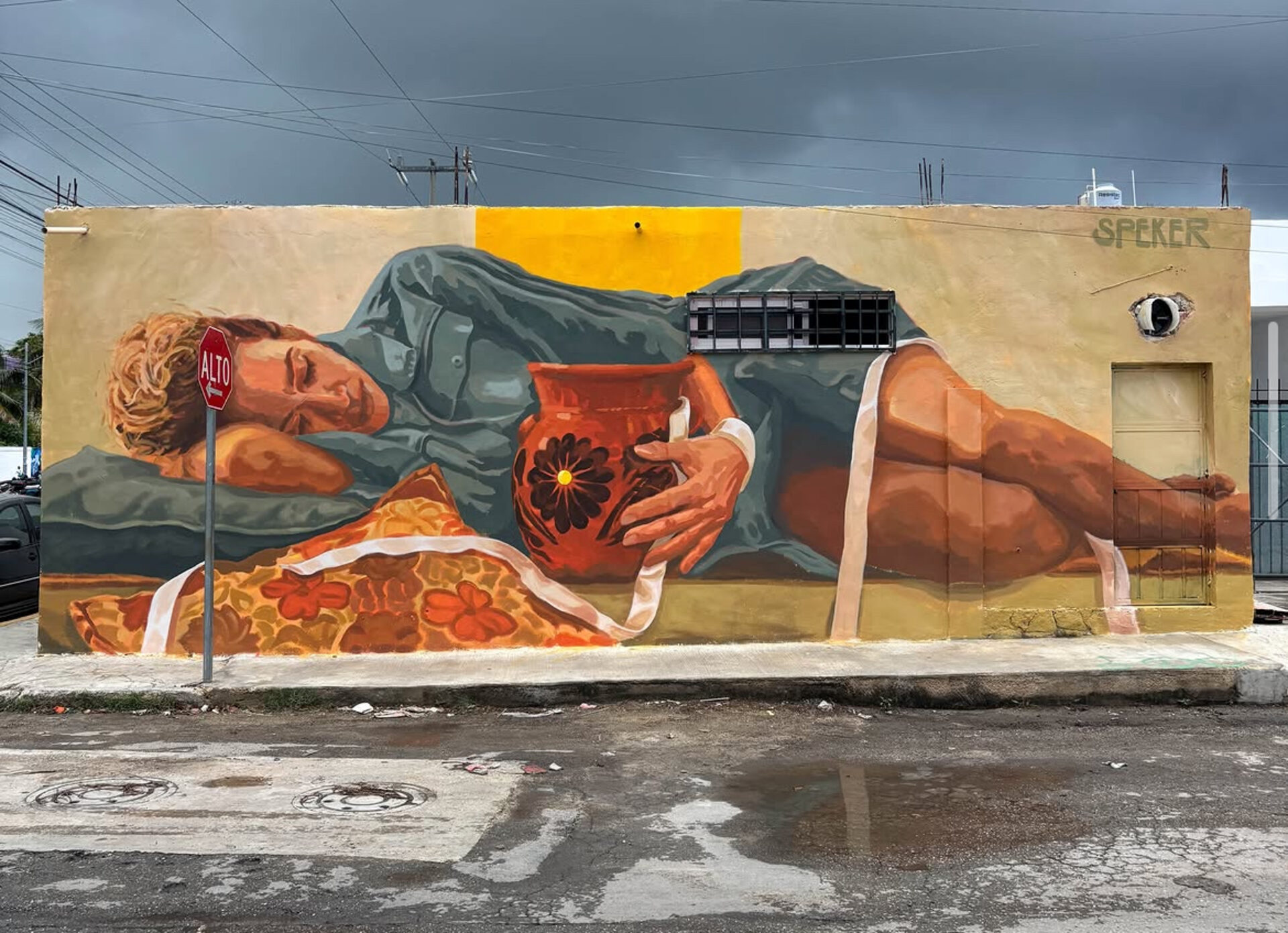 Large mural of a reclining figure holding a clay jug, painted across a street facing wall under a grey sky.